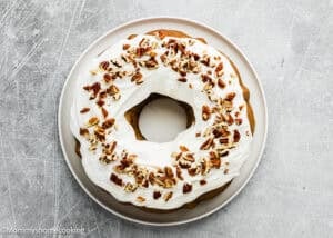 An Eggless Carrot Bundt Cake topped with white frosting and chopped pecans sits on a white plate against a light gray background.