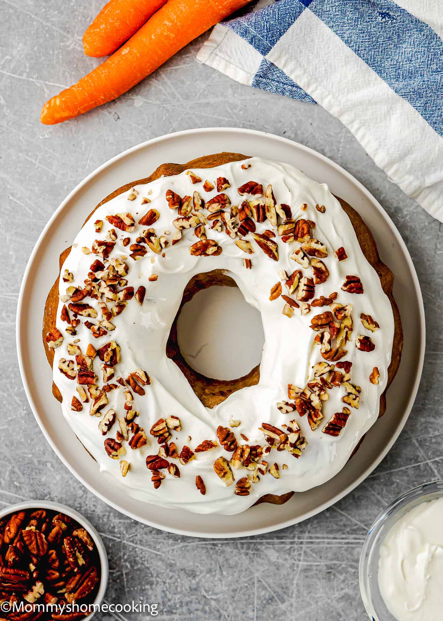 An Eggless Carrot Bundt Cake with white frosting and chopped pecans sits on a white plate. Nearby are carrots, a napkin, a bowl of pecans, and a bowl of frosting.