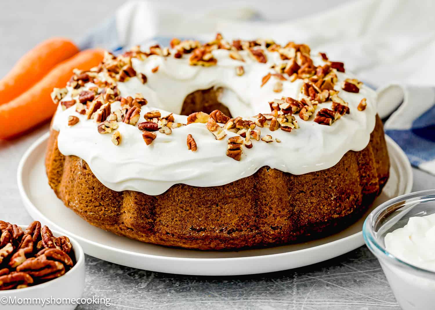 An Eggless Carrot Bundt Cake topped with white cream cheese frosting and chopped pecans sits on a white plate, with a bowl of pecans and carrots nearby.