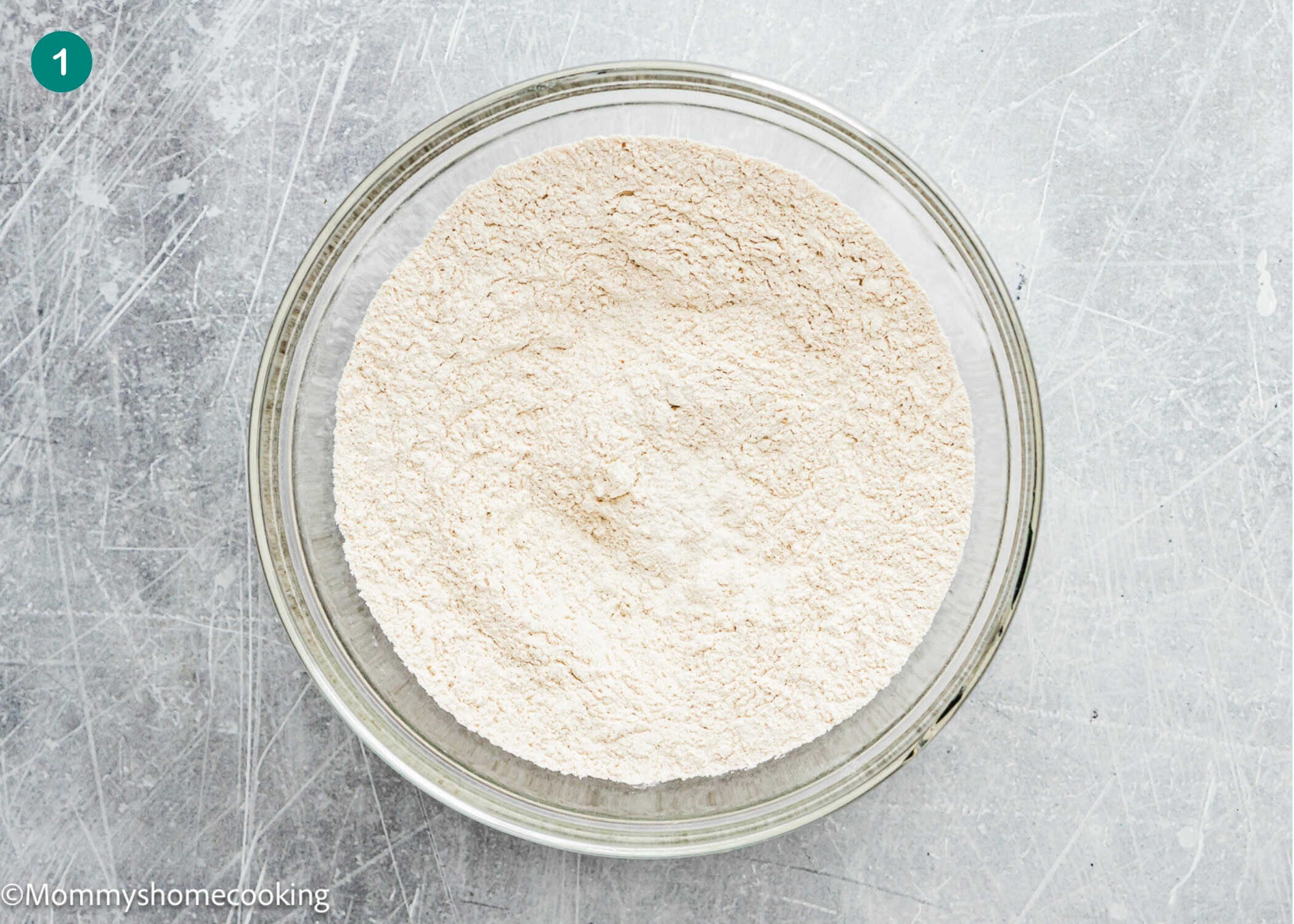 A glass bowl filled with mixed dry ingredients for Eggless Carrot Bundt Cake sits on a scratched gray surface.