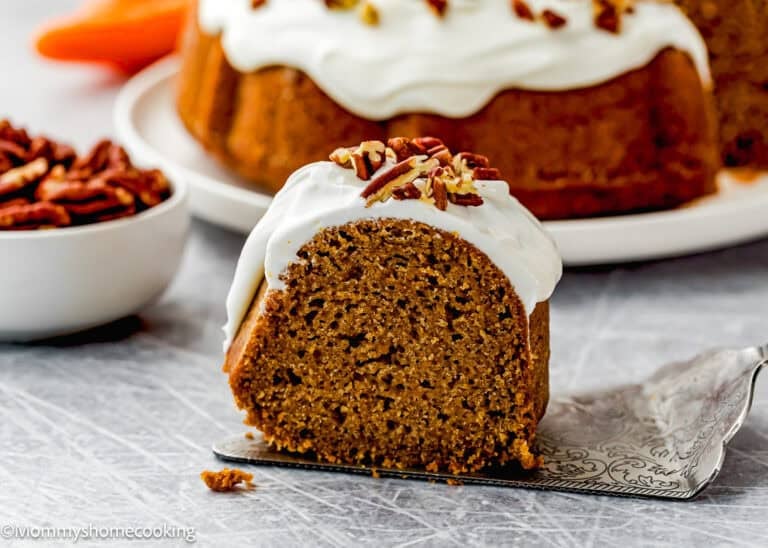 A slice of Eggless Carrot Bundt Cake with white frosting and chopped pecans on top, served on a metal cake server; whole cake and bowl of pecans in the background.