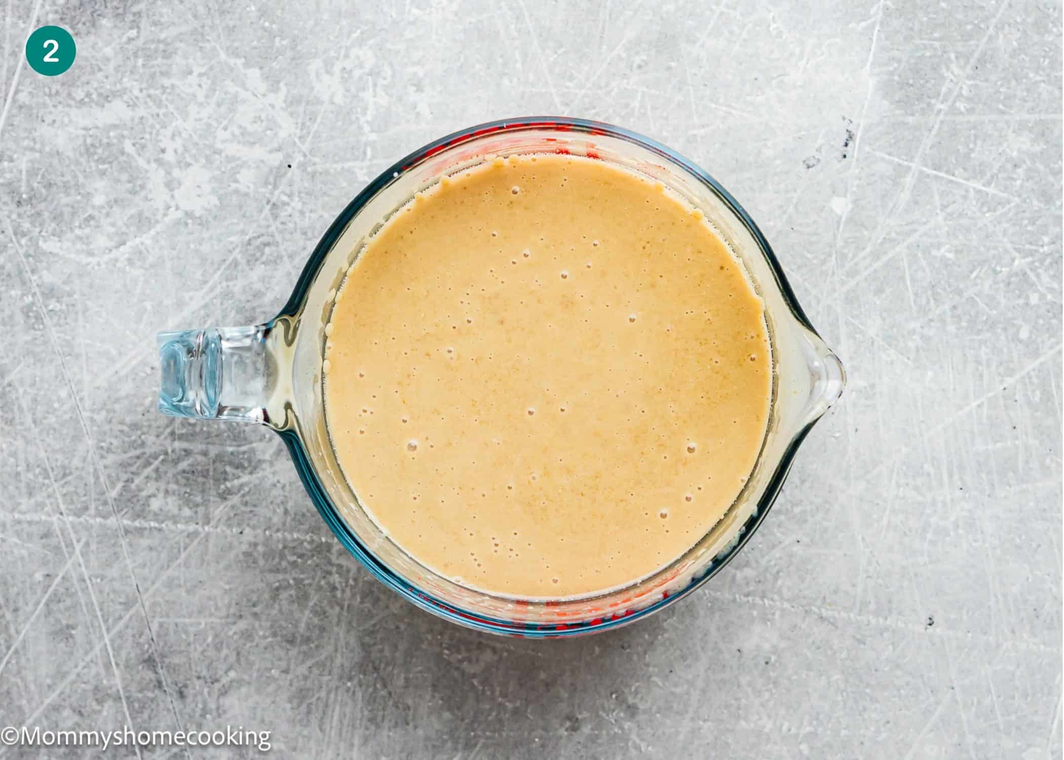 A glass measuring cup filled with a light brown liquid mixture for Eggless Carrot Bundt Cake sits on a gray textured surface.