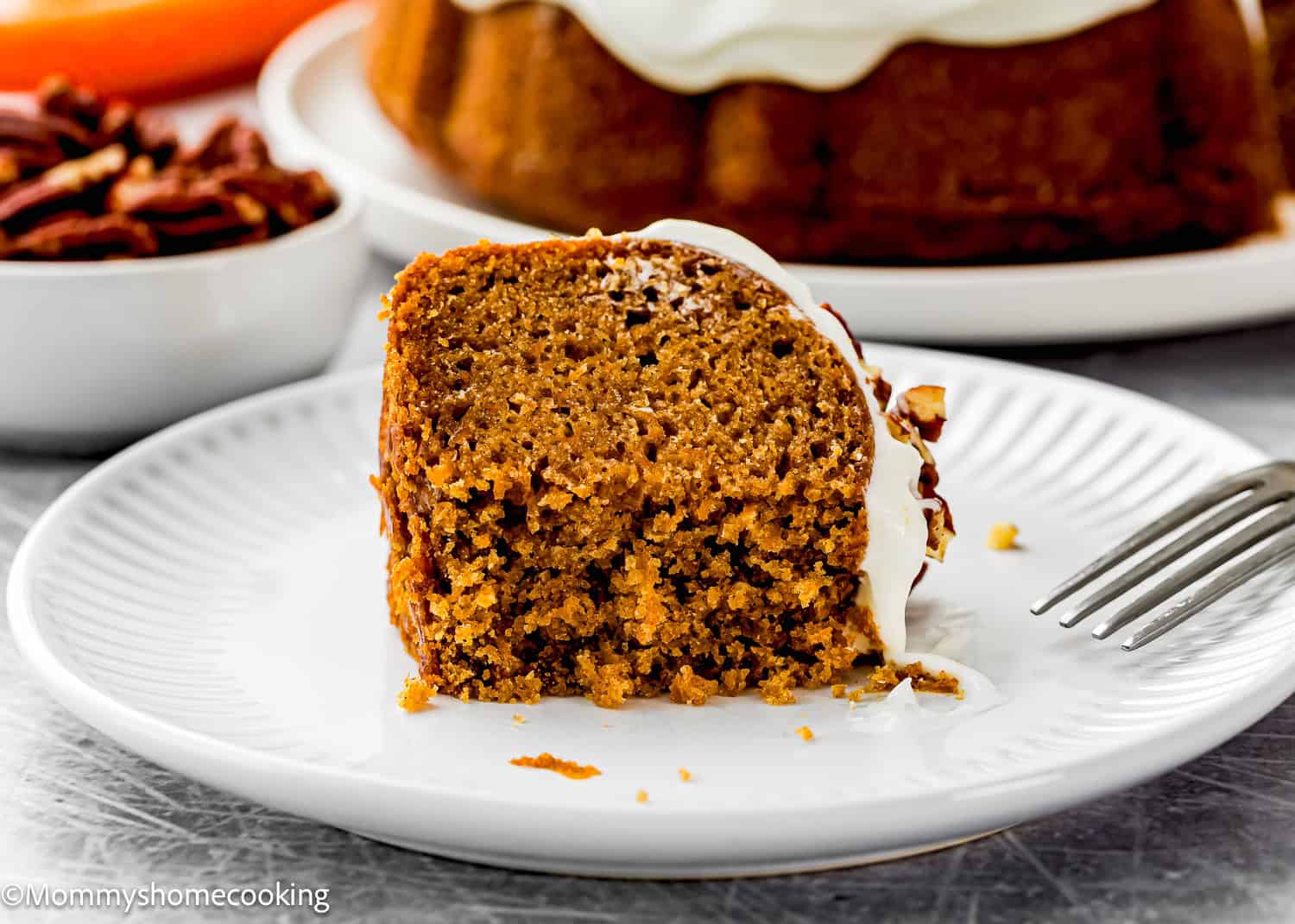 A slice of moist Eggless Carrot Bundt Cake with white frosting on a white plate, with part of the whole cake and a bowl of pecans in the background.