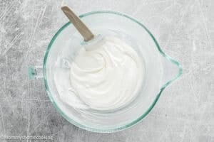 A glass mixing bowl with thick white whipped cream and a spatula, placed on a textured gray surface—perfect for preparing an Eggless Carrot Bundt Cake.