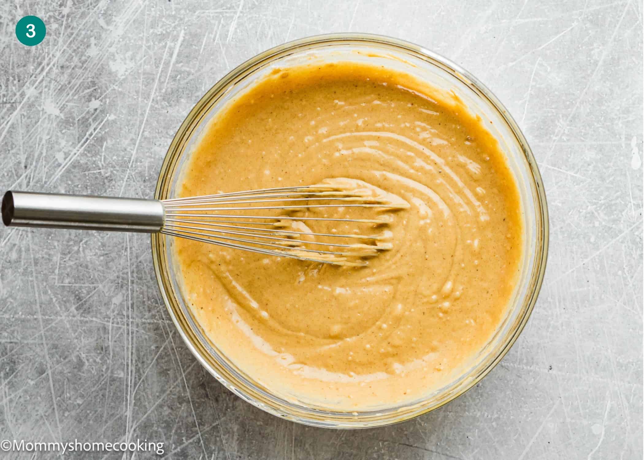 A metal whisk in a glass bowl of thick, light brown Eggless Carrot Bundt Cake batter on a scratched gray surface.