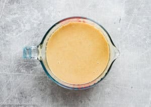 A glass measuring cup filled with a light brown mixture for Eggless Carrot Bundt Cake sits on a scratched gray surface.