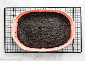 An Eggless Chocolate Brownie Pudding (Ina Garten–Inspired) sits in a rectangular pink baking dish, cooling on a black wire rack over a white surface.