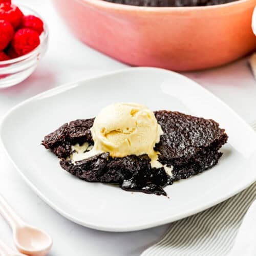 A serving of Eggless Chocolate Brownie Pudding (Ina Garten–Inspired) topped with a scoop of vanilla ice cream sits on a white plate, with a dish of raspberries and the remaining cake in the background.