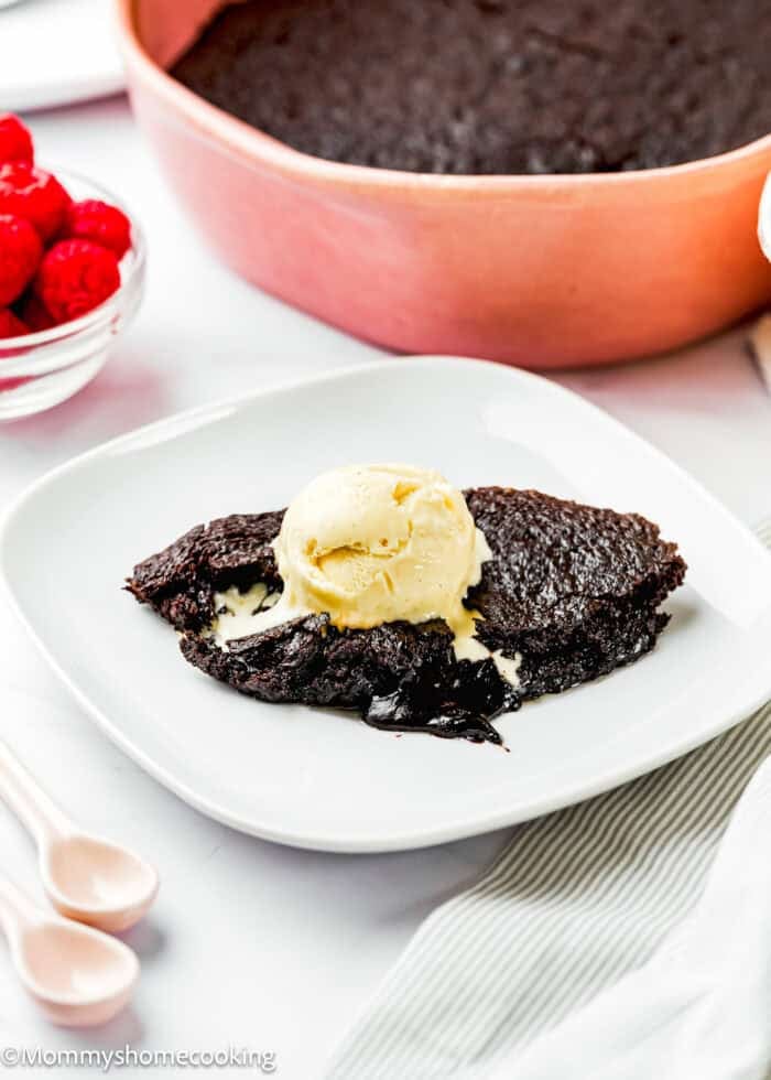 A serving of Eggless Chocolate Brownie Pudding (Ina Garten–Inspired) topped with a scoop of vanilla ice cream sits on a white plate, with a dish of raspberries and the remaining cake in the background.