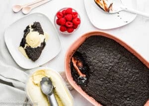 A serving of Eggless Chocolate Brownie Pudding with vanilla ice cream on a plate, a bowl of raspberries, a baking dish with more pudding, and a container of ice cream with a scoop.