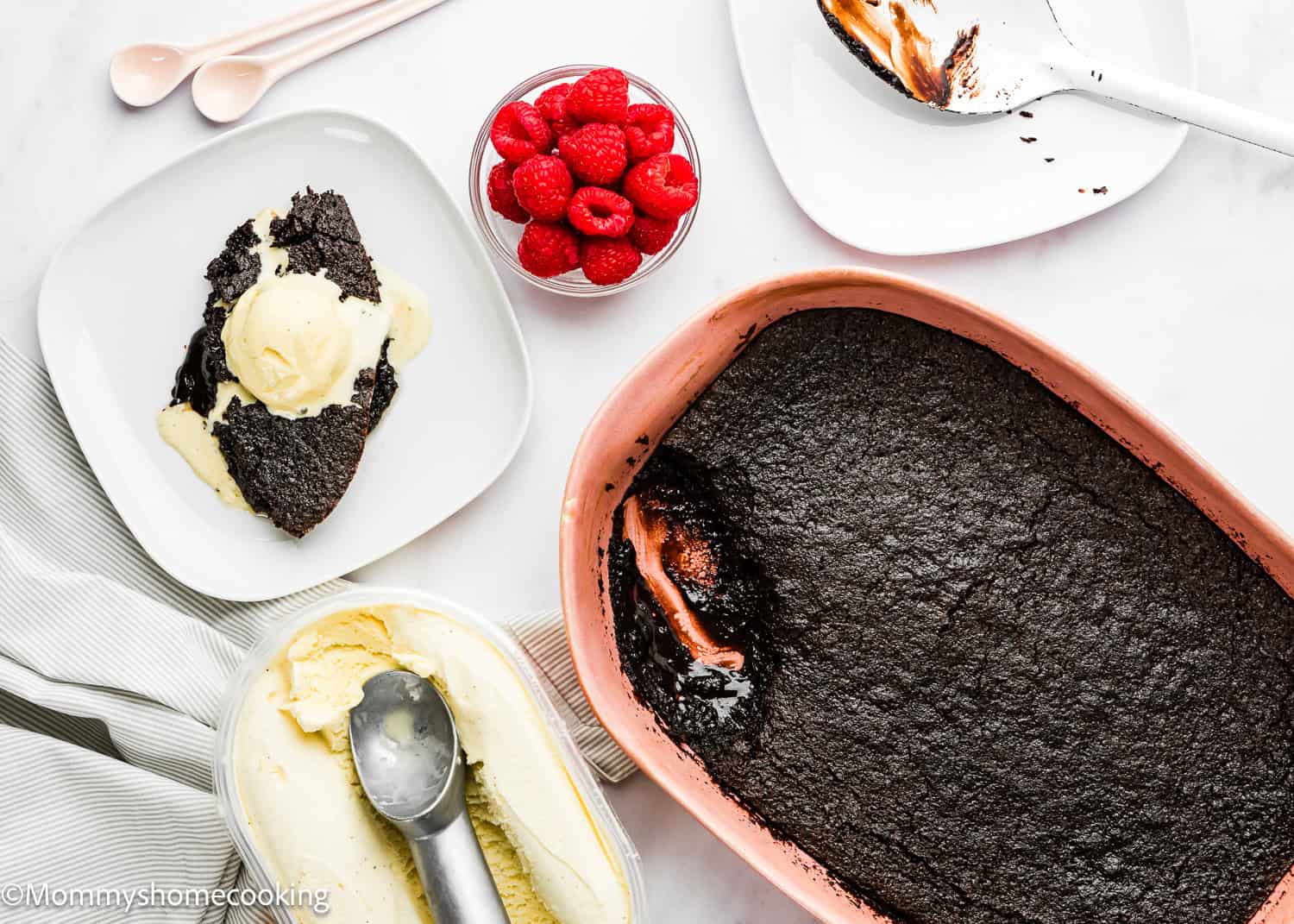 A serving of Eggless Chocolate Brownie Pudding with vanilla ice cream on a plate, a bowl of raspberries, a baking dish with more pudding, and a container of ice cream with a scoop.