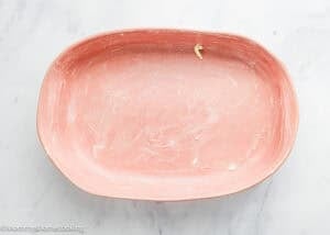 An empty, pink baking dish with remnants of butter or grease rests on a white marble countertop, hinting at freshly baked Eggless Chocolate Brownie Pudding (Ina Garten–Inspired).