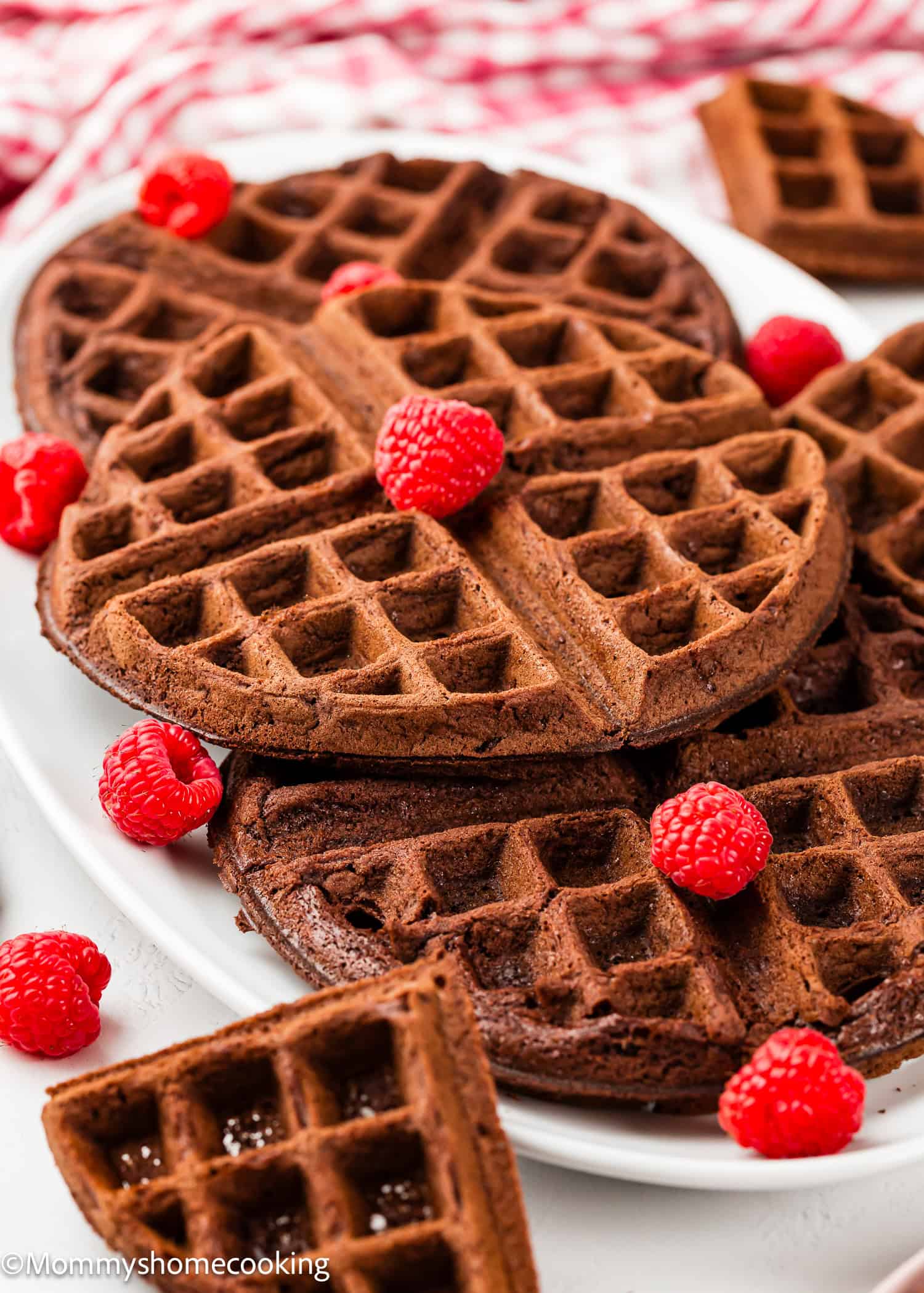 A plate of eggless chocolate waffles garnished with fresh raspberries, with more waffles and raspberries arranged around the plate on a white surface.