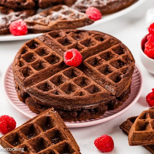 A stack of Eggless Chocolate Waffles on a pink plate, topped with a raspberry, surrounded by loose raspberries and waffle pieces, with a bowl of raspberries nearby.