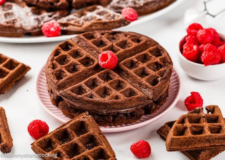 A stack of Eggless Chocolate Waffles on a pink plate, topped with a raspberry, surrounded by loose raspberries and waffle pieces, with a bowl of raspberries nearby.