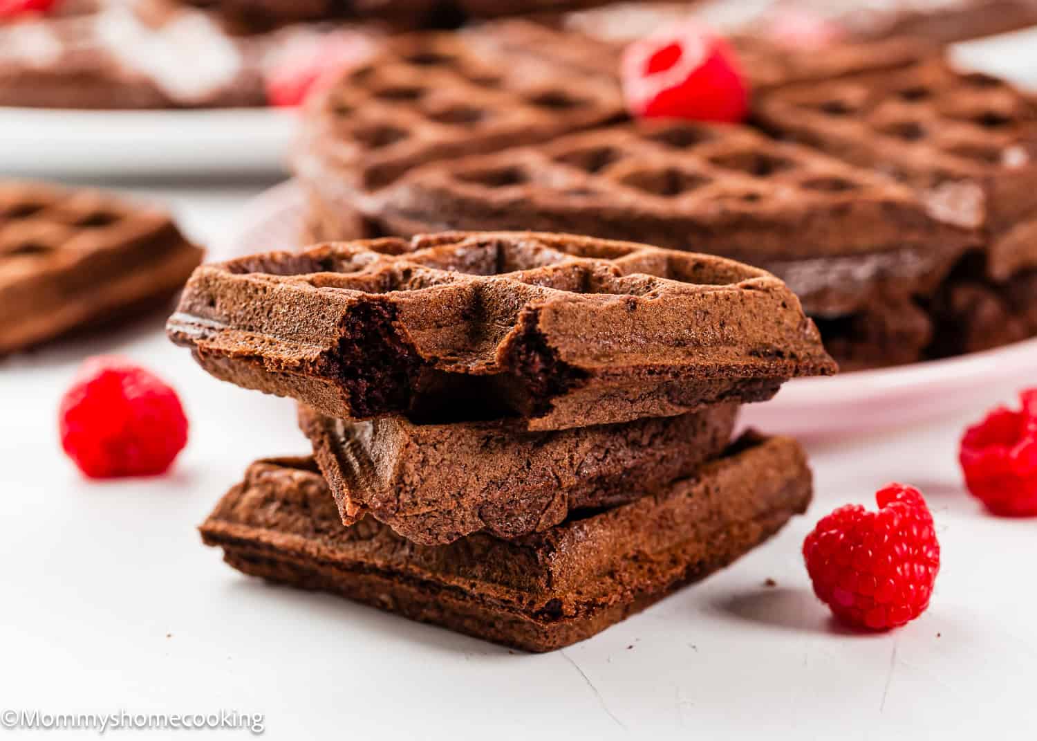Three stacked eggless chocolate waffles with visible bite marks, surrounded by fresh raspberries, with more waffles in the background.