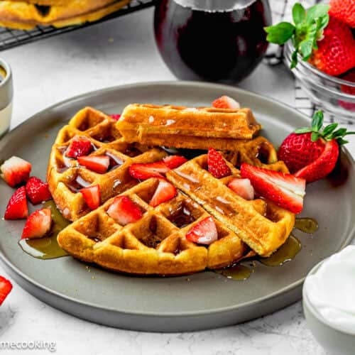 A plate of Eggless Yogurt Waffles topped with sliced strawberries and syrup, served with a side of whipped cream, fresh strawberries, and extra syrup on a white table.