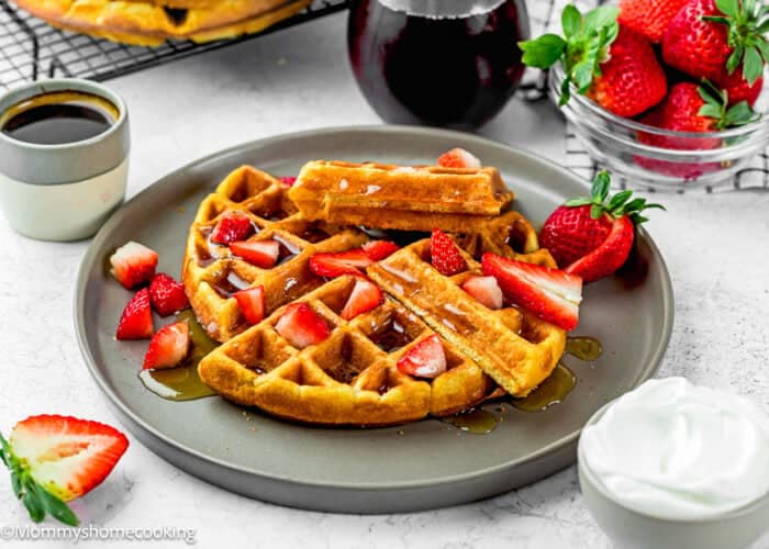 A plate of Eggless Yogurt Waffles topped with sliced strawberries and syrup, served with a side of whipped cream, fresh strawberries, and extra syrup on a white table.