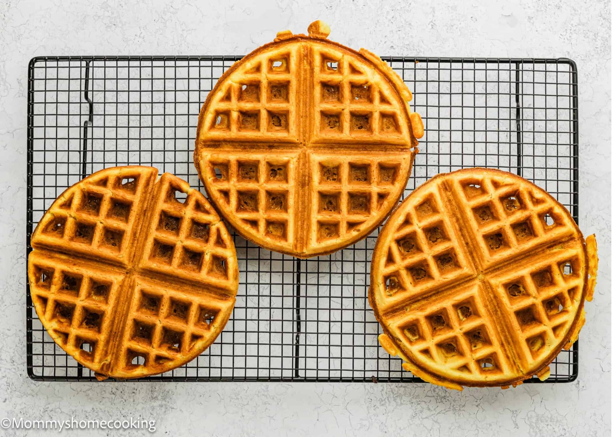 Three round, golden Eggless Yogurt Waffles cool on a black wire rack set on a light, textured surface.
