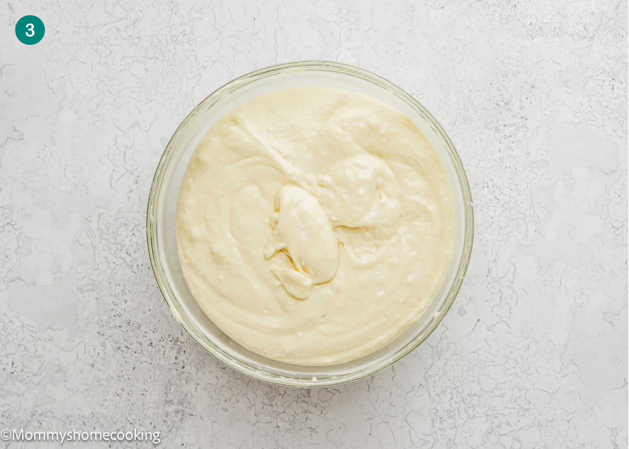 A glass bowl filled with smooth, creamy batter for Eggless Yogurt Waffles sits on a light, textured surface.