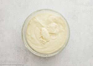 A glass bowl filled with smooth, creamy cake batter for Eggless Yogurt Waffles sits on a light, textured surface.
