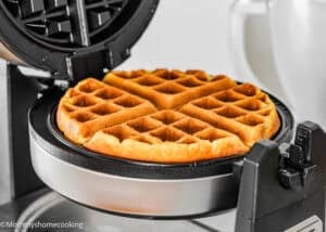 An eggless yogurt waffle cooks in an open electric waffle maker, with a white mixing bowl in the background.