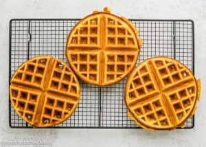 Three golden-brown Eggless Yogurt Waffles cool on a black wire rack placed on a light-colored surface.