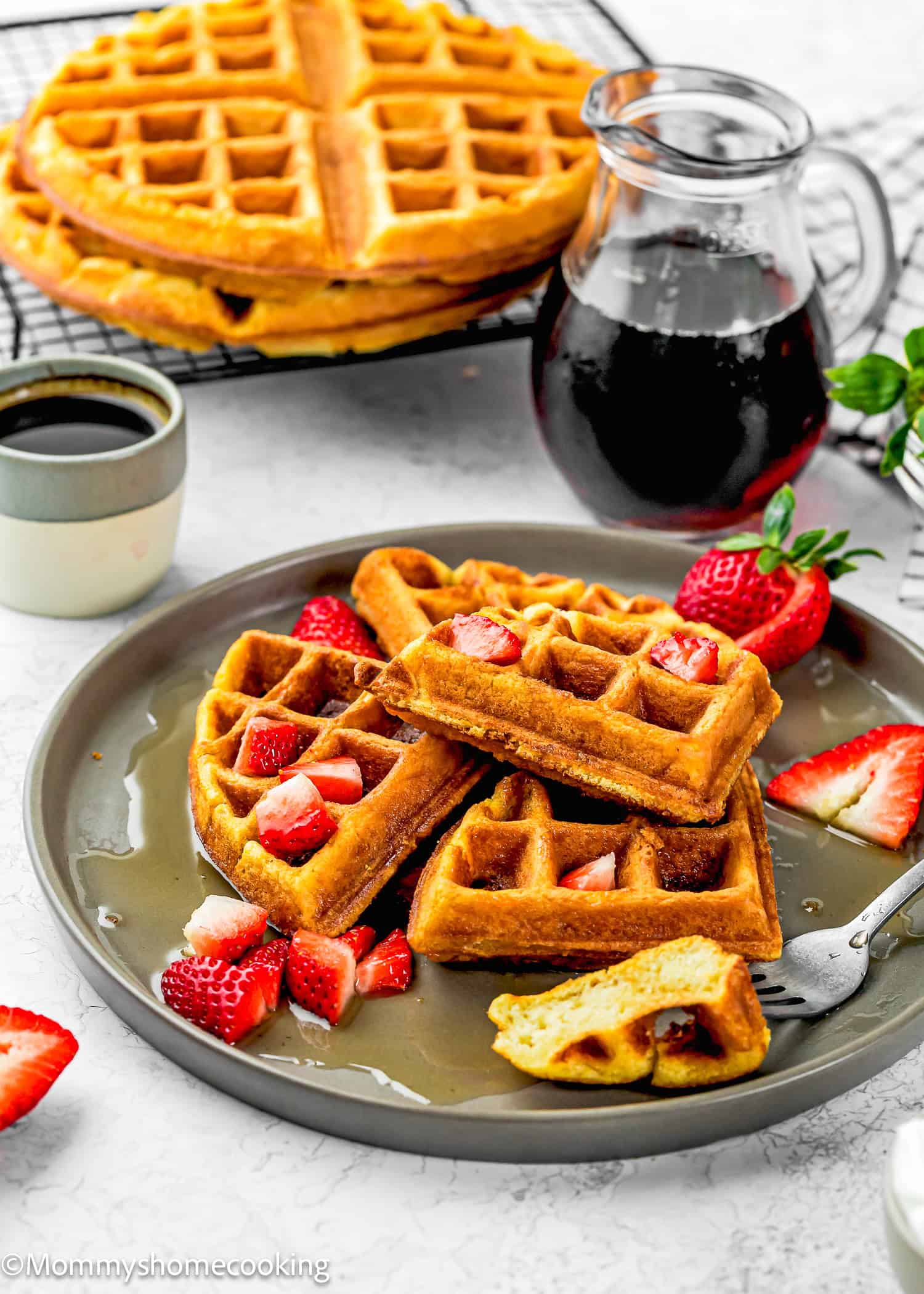 A plate of Eggless Yogurt Waffles topped with strawberries and syrup, a fork, a small cup, a jug of syrup, and whole waffles on a cooling rack in the background.