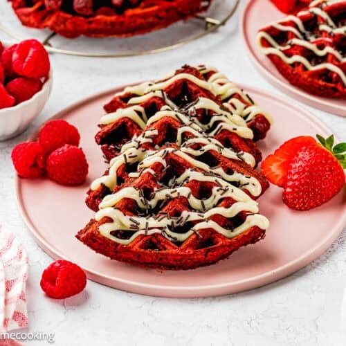 A plate of Fluffy Red Velvet Waffles (No Eggs) drizzled with white icing, topped with sprinkles, and served with fresh raspberries and a strawberry on the side.