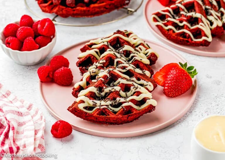 A plate of Fluffy Red Velvet Waffles (No Eggs) drizzled with white icing, topped with sprinkles, and served with fresh raspberries and a strawberry on the side.