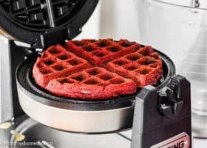 A fluffy red velvet waffle (no eggs) is baking in an open waffle maker, with a clear mixing bowl in the background.