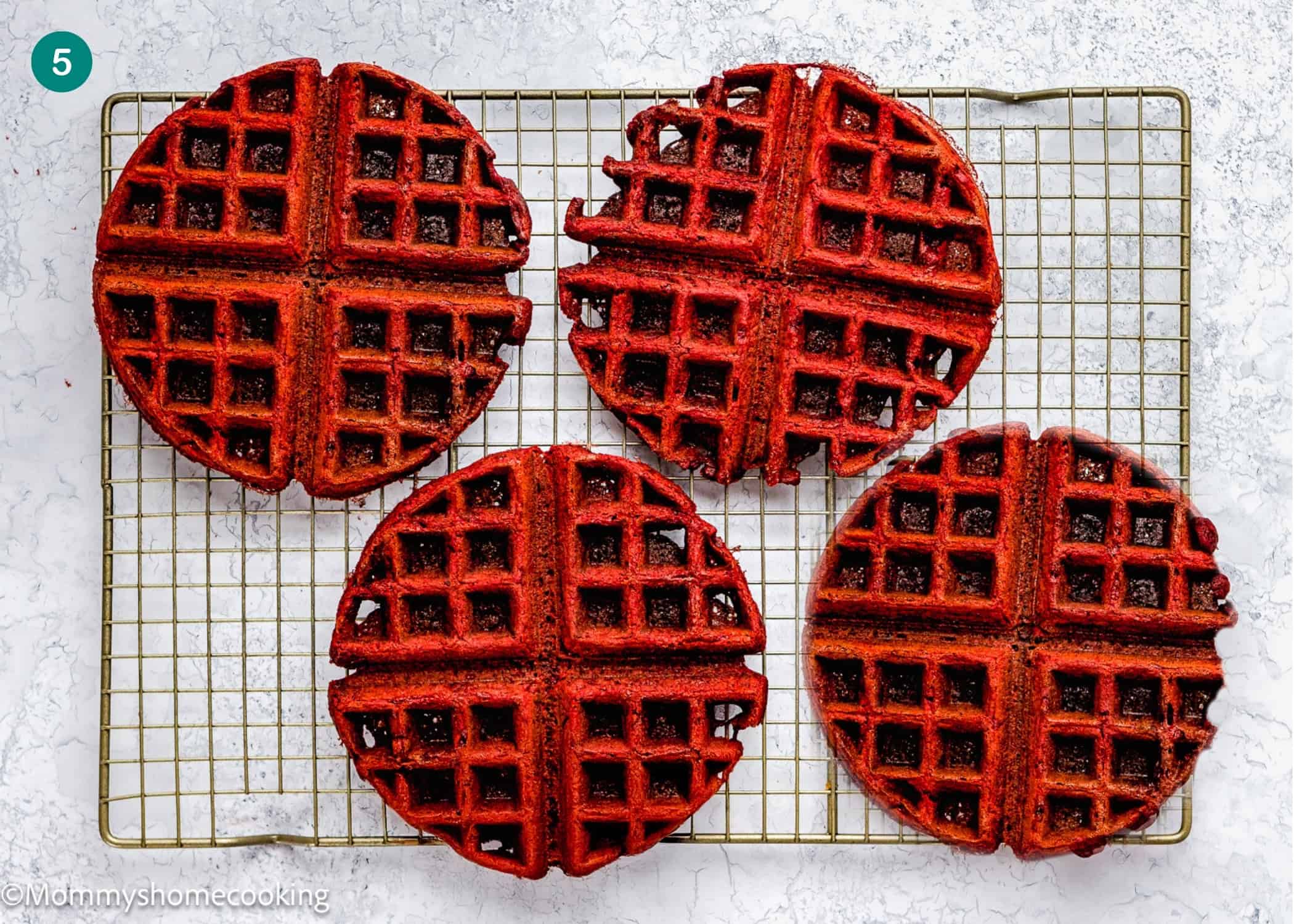 Four fluffy Red Velvet Waffles (No Eggs) are cooling on a wire rack placed on a white textured surface.
