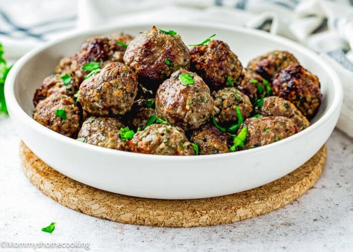 A white bowl filled with cooked meatballs from a simple meatball recipe without eggs, garnished with chopped parsley, sits on a cork trivet.