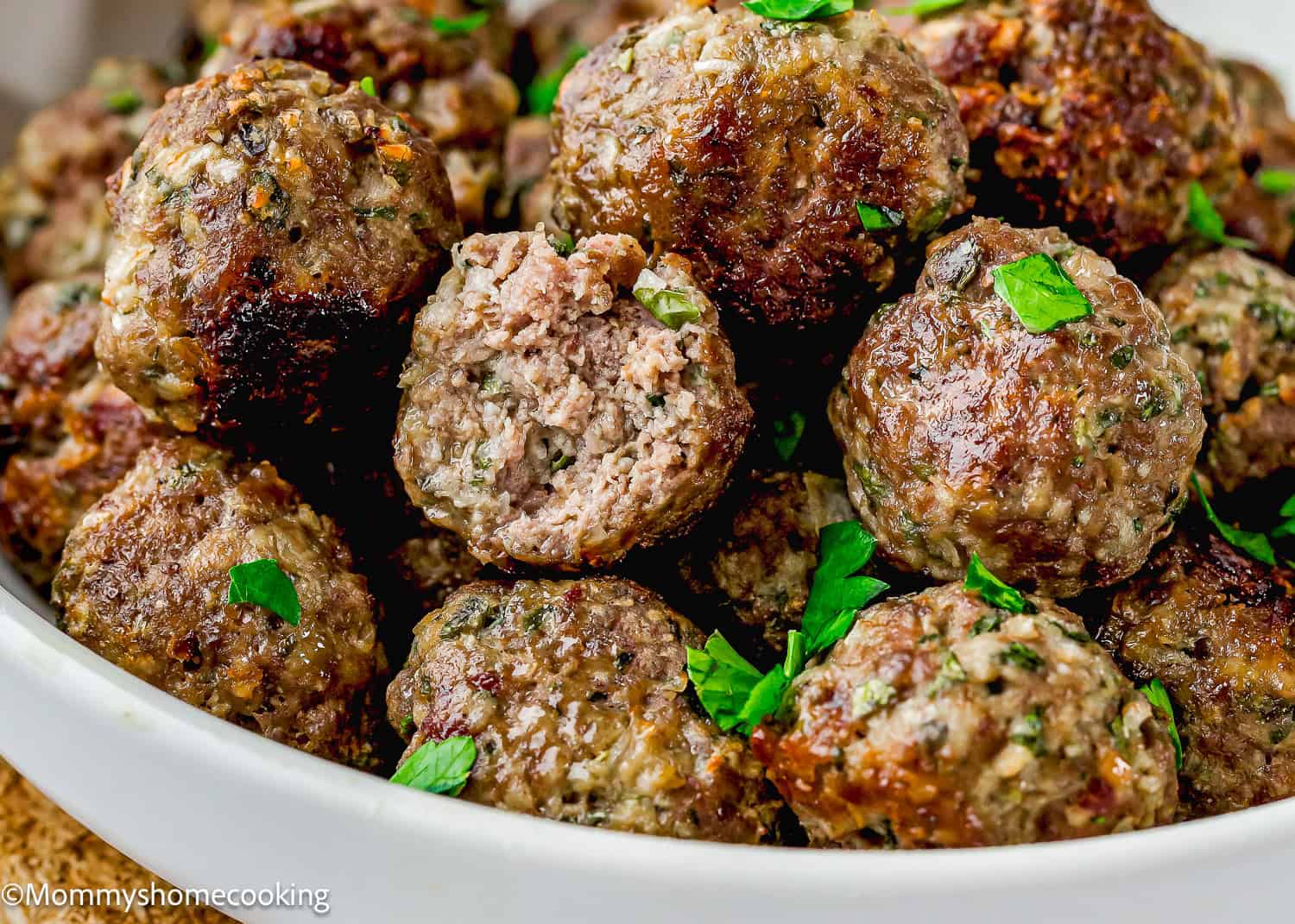 A close-up of cooked meatballs garnished with chopped parsley, piled in a white bowl. One meatball is sliced open to reveal the inside texture—perfect for a Simple Meatball Recipe Without Eggs.