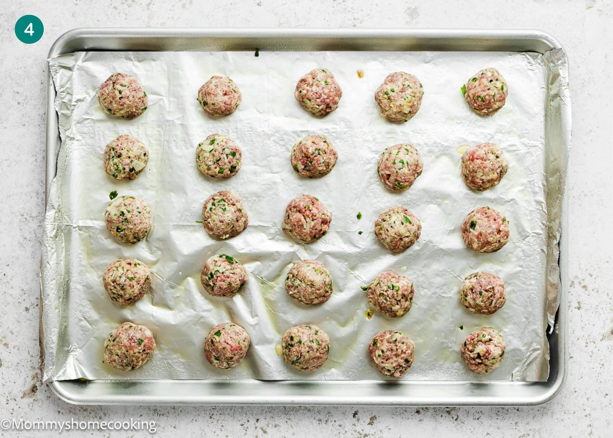 A baking sheet lined with foil holds 24 uncooked meatballs arranged in rows, ready to bake using a Simple Meatball Recipe Without Eggs.