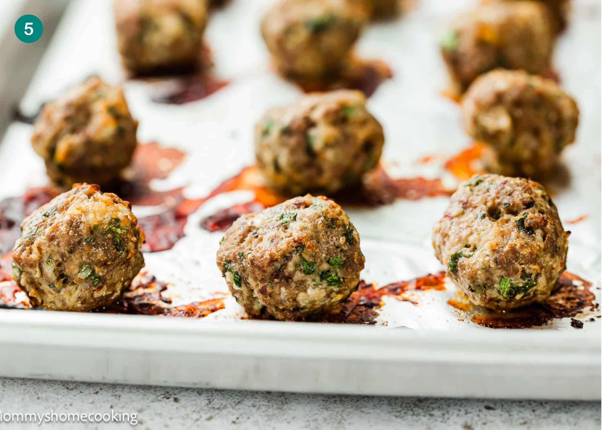Several cooked meatballs with herbs are arranged on a parchment-lined baking sheet, with browned spots from roasting visible underneath—a simple meatball recipe without eggs.