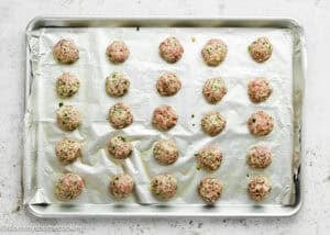 A baking sheet lined with foil holds 25 evenly spaced, uncooked meatballs mixed with herbs and vegetables—perfect for a simple meatball recipe without eggs, ready to be baked.
