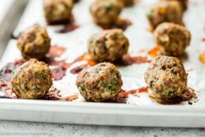A baking sheet with several cooked meatballs resting on parchment paper, showing browned spots and bits of herbs from a simple meatball recipe without eggs.