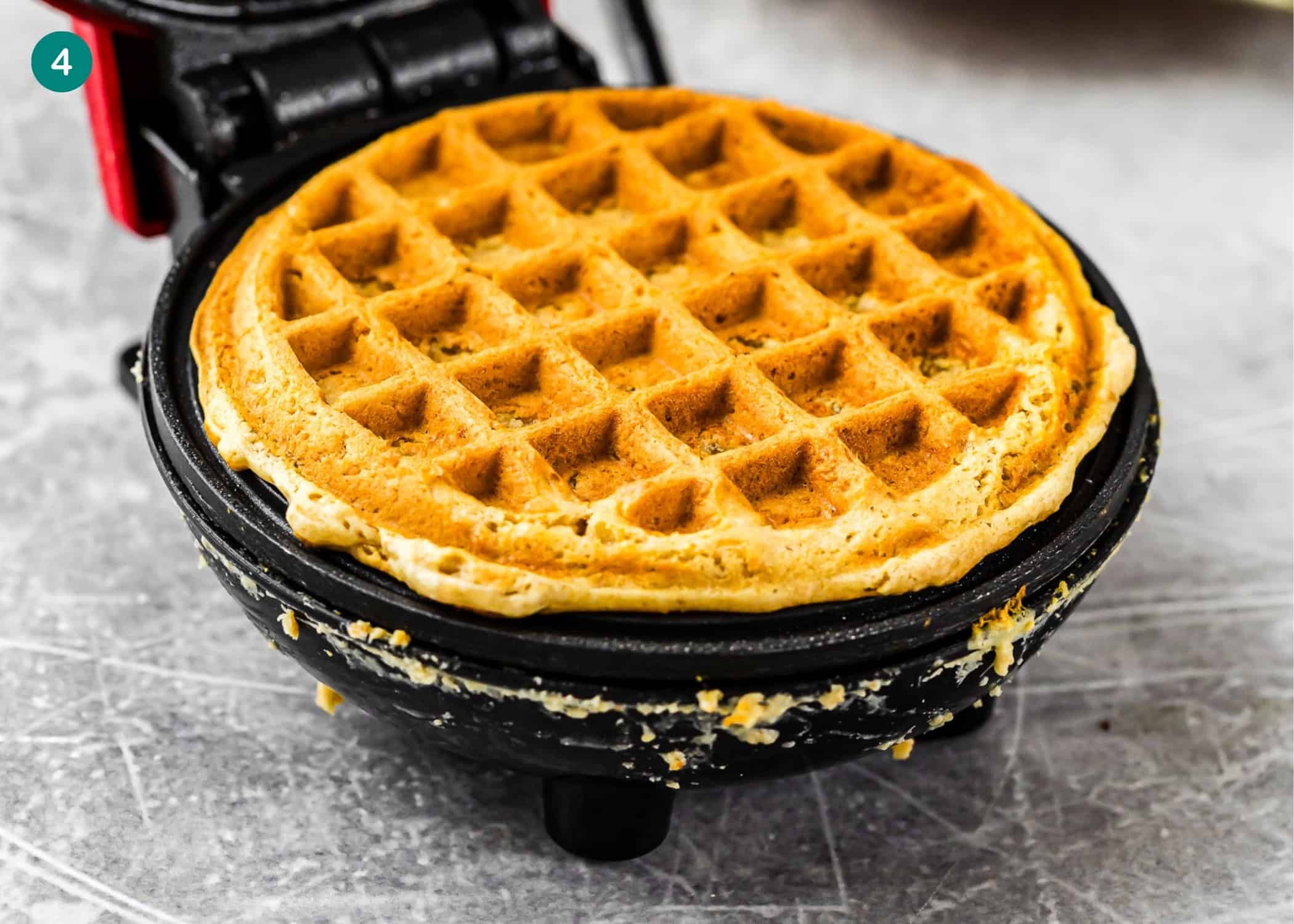 A freshly cooked golden-brown egg-free, dairy-free, and refined sugar–free oat waffle sits in an open waffle maker with some batter residue around the edges.