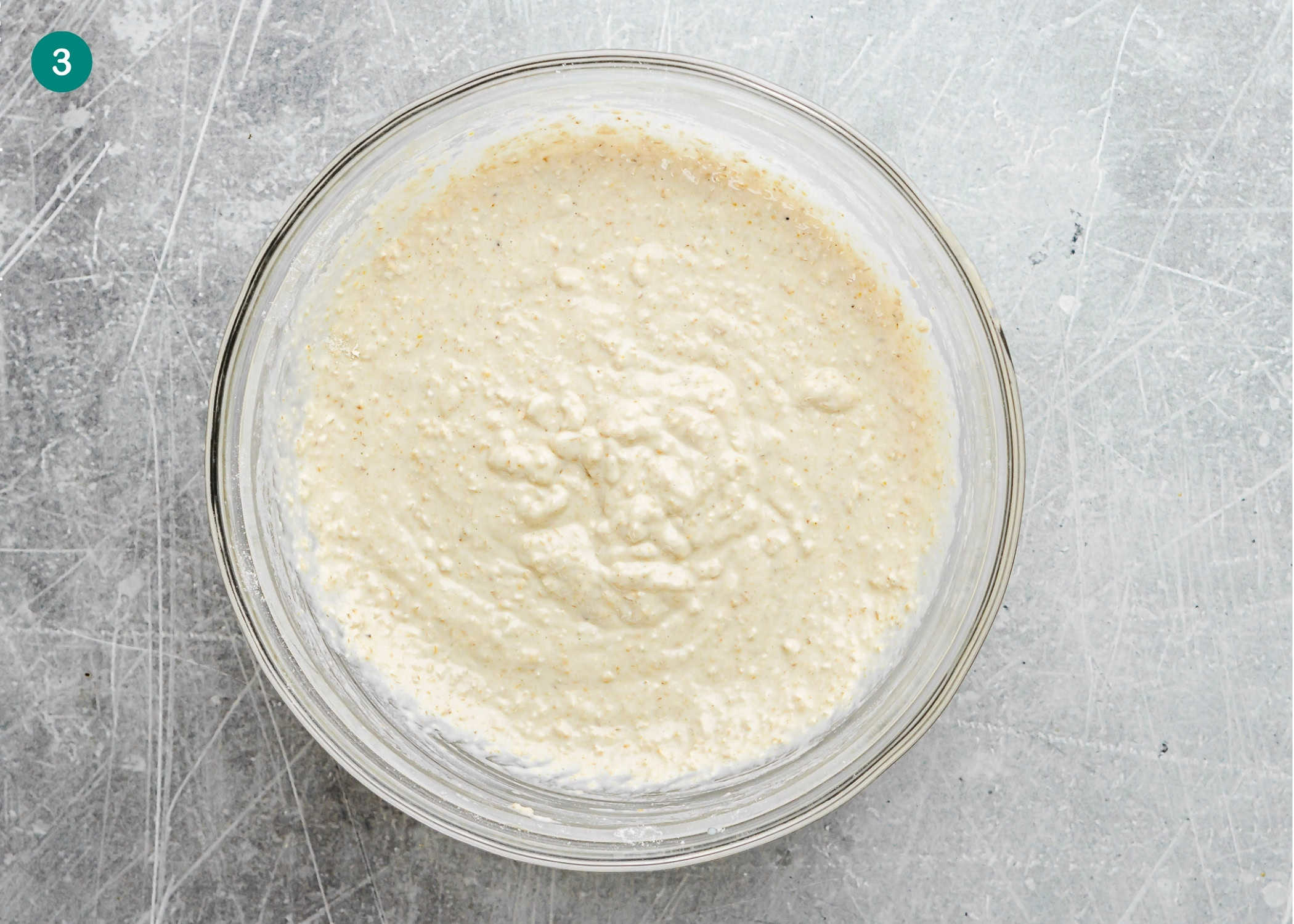 A glass bowl filled with thick, slightly lumpy batter for dairy-free and refined sugar–free oat waffles sits on a scratched, light gray surface.