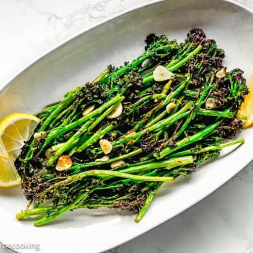 Oval plate with Air Fryer Broccolini with Garlic, sliced garlic, and lemon wedges on a white marble surface.