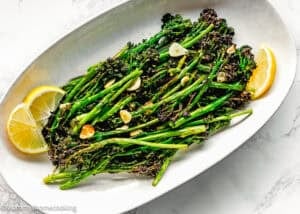 A white oval plate with Air Fryer Broccolini with Garlic, topped with sliced garlic and lemon wedges on the side.