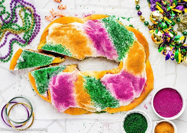 A sliced Best Eggless Mardi Gras King Cake decorated with green, purple, and gold sugar sits on a white surface, surrounded by Mardi Gras beads and decorations.