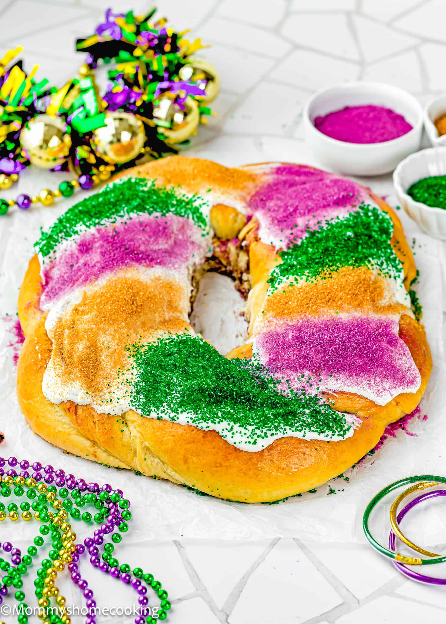 A round Best Eggless Mardi Gras King Cake decorated with purple, green, and yellow sugar sits on a white surface, surrounded by Mardi Gras beads, masks, and bowls of colored sugar.