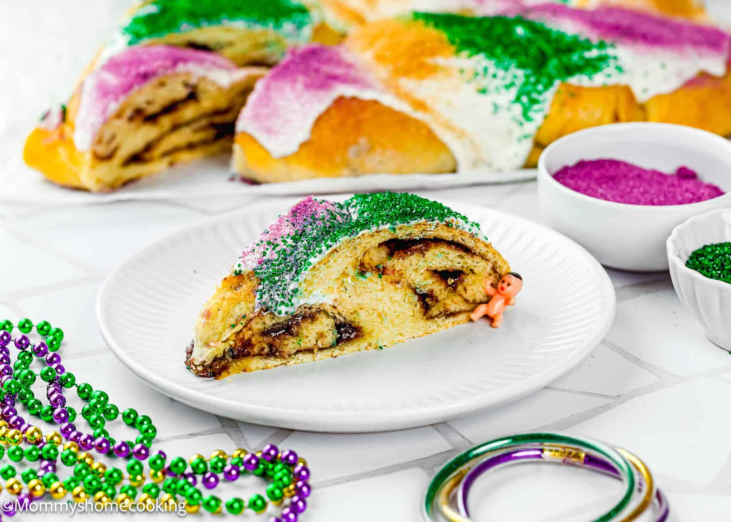 A slice of the Best Eggless Mardi Gras King Cake topped with green, purple, and yellow sugar sits on a white plate, with a small plastic baby beside it, beads, and colored sugar bowls nearby.