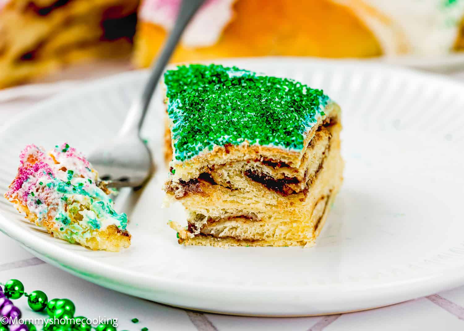 A slice of Best Eggless Mardi Gras King Cake with green sugar topping sits on a white plate beside a fork and a bite-sized piece of cake with purple sprinkles.