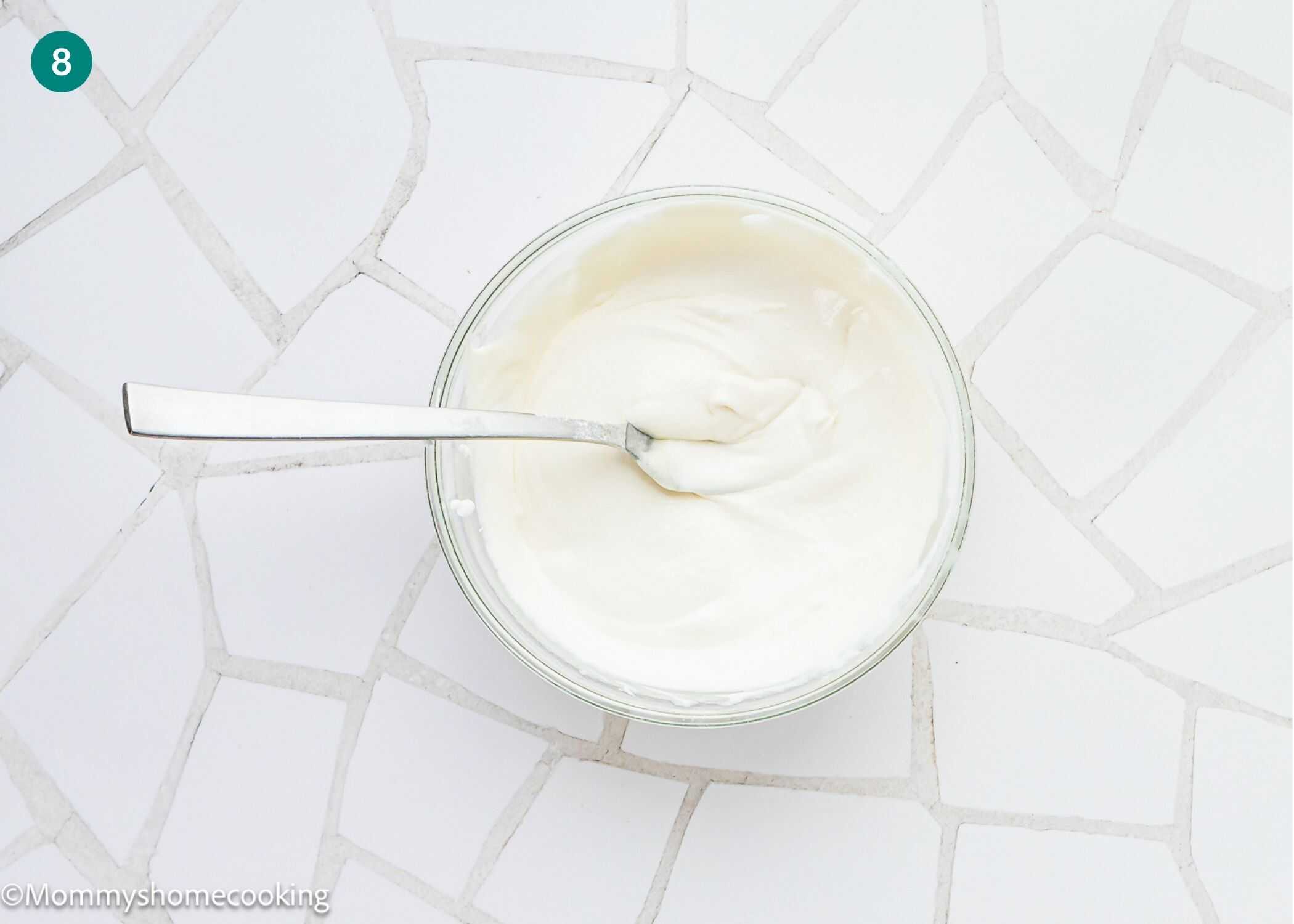 A glass bowl filled with a smooth white creamy mixture and a spoon, placed on a white tile surface—perfect for topping the Best Eggless Mardi Gras King Cake.
