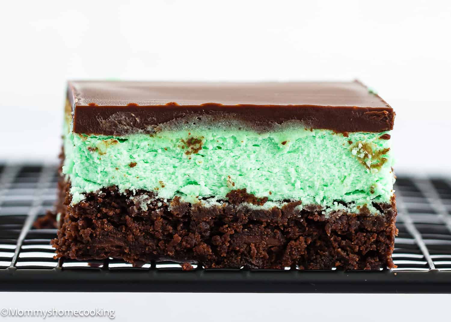 A close-up of Classic Eggless Mint Chocolate Brownies with a brownie base, green mint layer, and chocolate topping, resting on a black cooling rack.