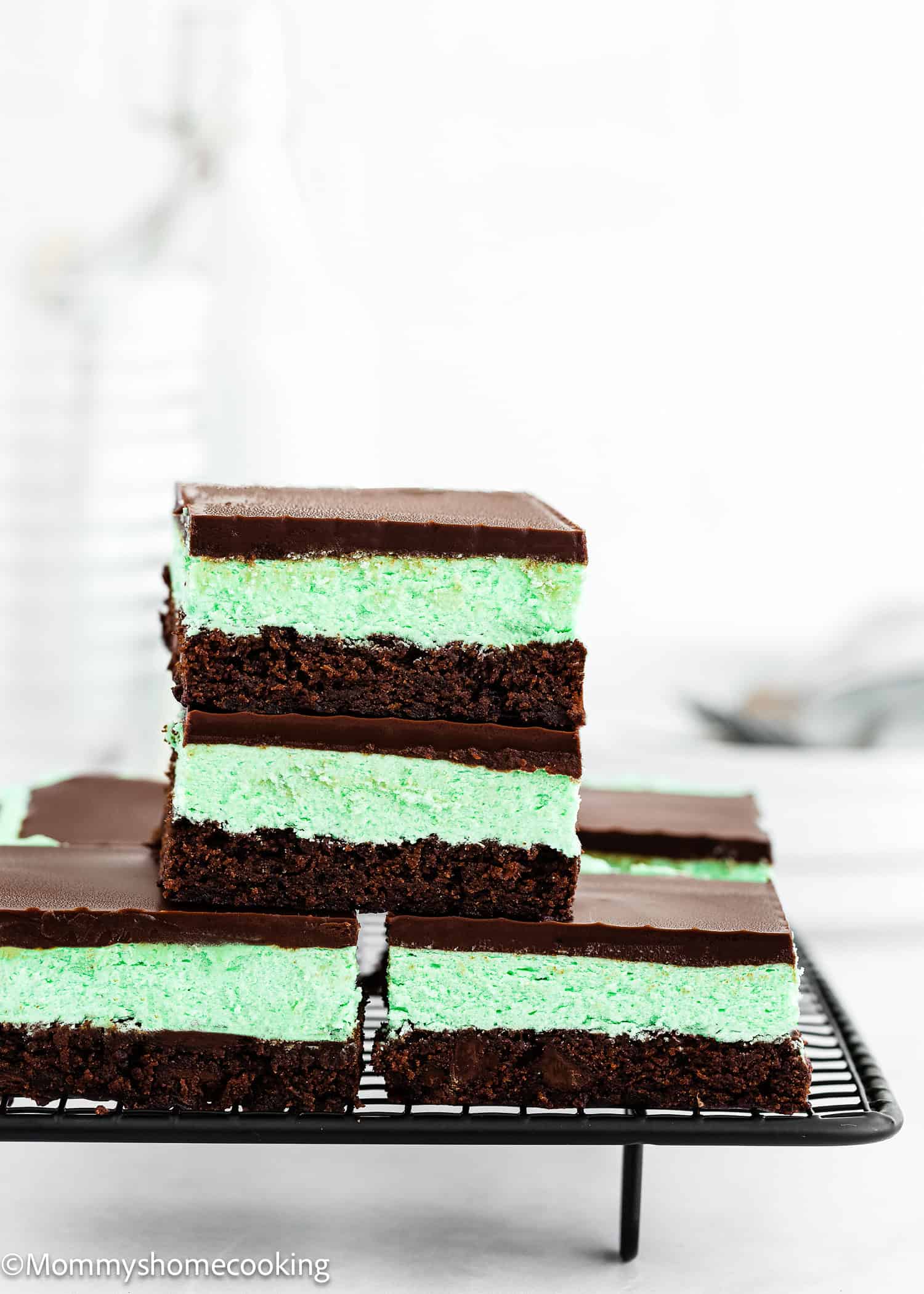 Three Classic Eggless Mint Chocolate Brownies are stacked on a cooling rack, featuring a fudgy chocolate base, green mint layer, and rich chocolate topping. More bars are visible in the background.