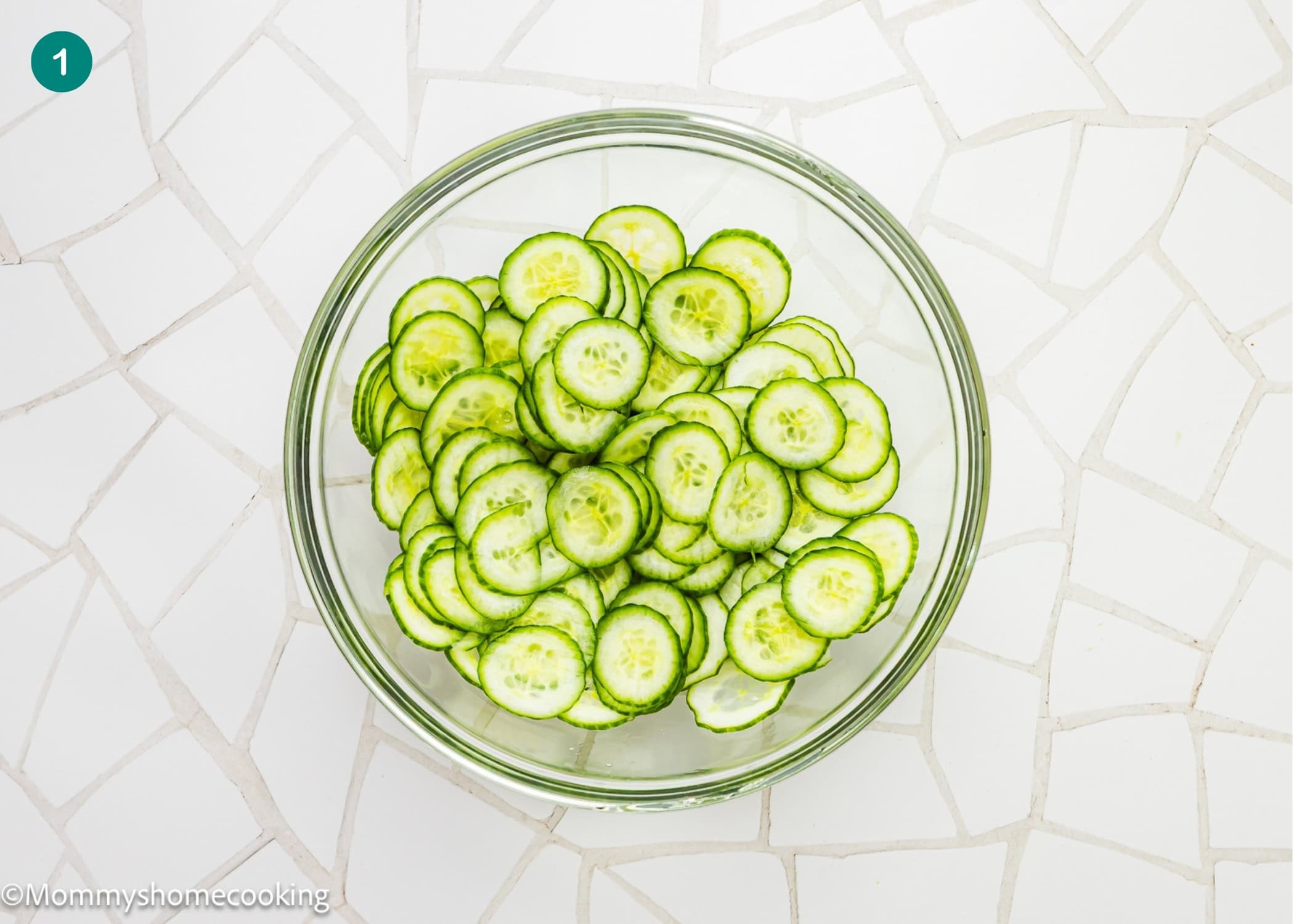 A clear glass bowl filled with thinly sliced cucumbers sits on a white tiled surface, ready to be made into a creamy cucumber salad (egg-free).